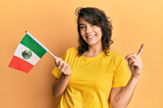 Foto jovem mulher hispânica segurando a bandeira do méxico sorrindo feliz apontando com a mão e o dedo para o lado