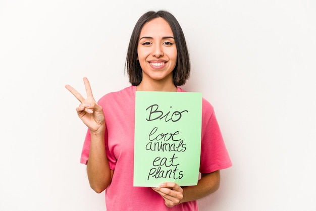 Foto jovem mulher hispânica segurando bio cartaz isolado no fundo branco alegre e despreocupado, mostrando um símbolo de paz com os dedos