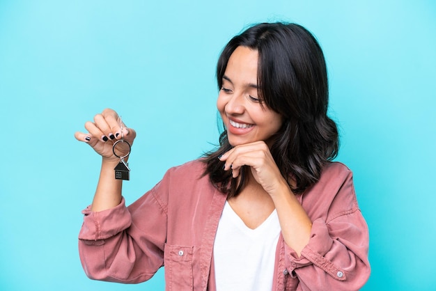 Foto jovem mulher hispânica segurando chaves de casa isolada em fundo azul olhando para o lado e sorrindo