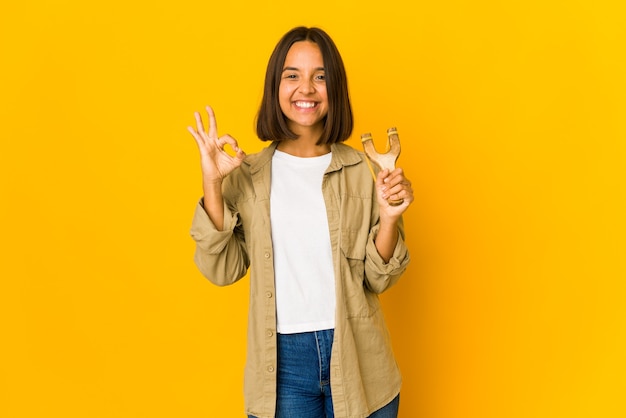 Jovem mulher hispânica segurando um estilingue alegre e confiante, mostrando o gesto de ok.