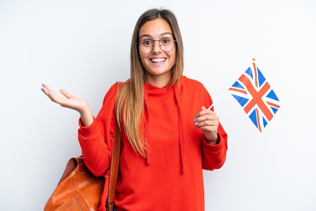 Jovem mulher hispânica segurando uma bandeira do Reino Unido isolada no fundo branco com expressão facial chocada