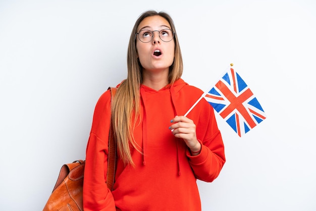 Jovem mulher hispânica segurando uma bandeira do Reino Unido isolada no fundo branco, olhando para cima e com expressão de surpresa