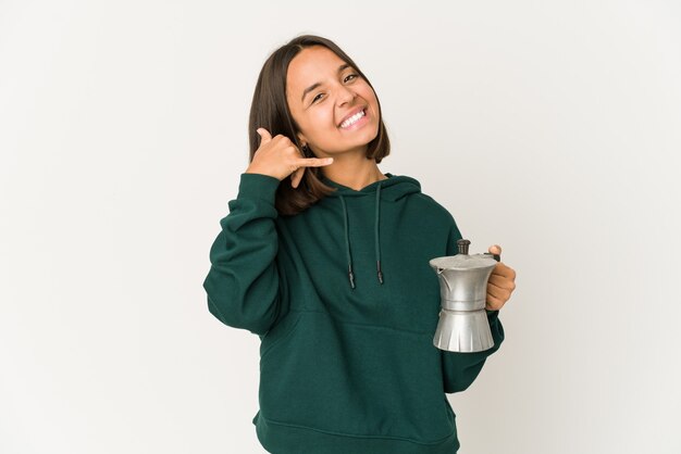 Jovem mulher hispânica segurando uma cafeteira, mostrando um gesto de chamada de celular com os dedos.