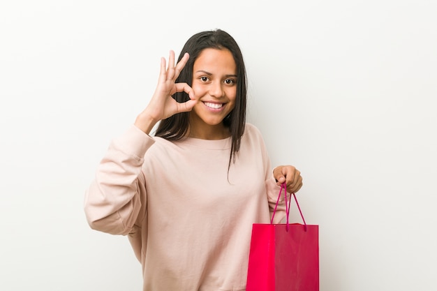 Jovem mulher hispânica segurando uma sacola de compras alegre e confiante, mostrando um gesto de ok.