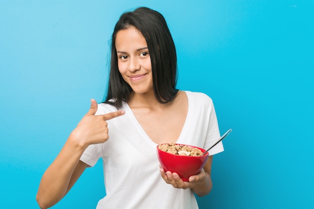 Jovem mulher hispânica, segurando uma tigela de cereal, apontando com o dedo para você, como se estivesse convidando para se aproximar.