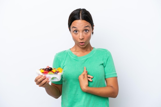 Jovem mulher hispânica segurando uma tigela de frutas isolada no fundo branco com expressão facial de surpresa