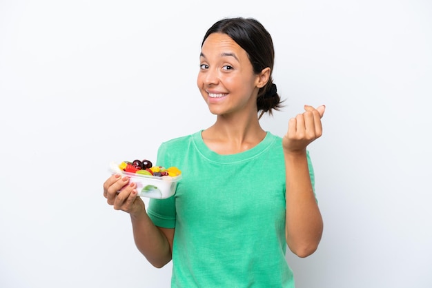 Jovem mulher hispânica segurando uma tigela de frutas isolada no fundo branco fazendo gesto de dinheiro