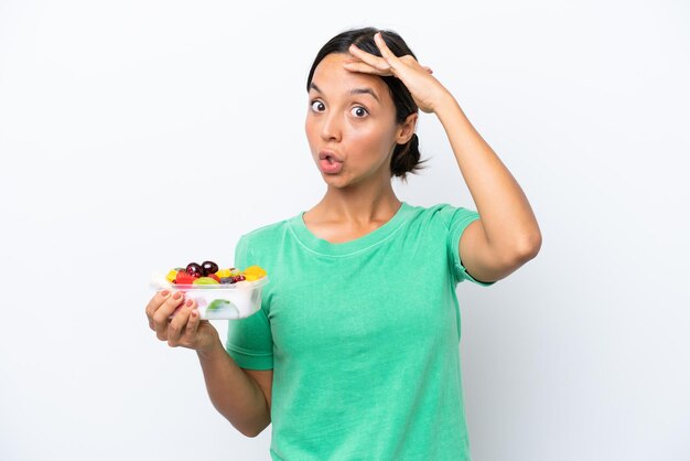 Jovem mulher hispânica segurando uma tigela de frutas isolada no fundo branco fazendo gesto de surpresa enquanto olha para o lado
