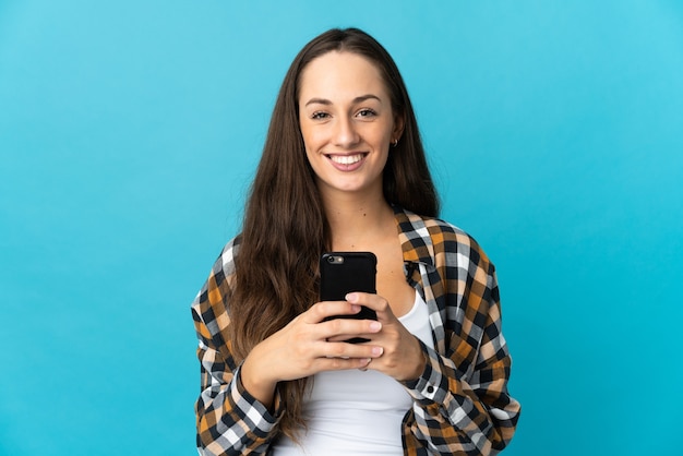 Foto jovem mulher hispânica sobre fundo azul isolado, olhando para a câmera e sorrindo enquanto usa o celular