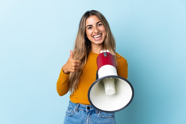 Jovem mulher hispânica sobre uma parede azul isolada segurando um megafone com o polegar para cima