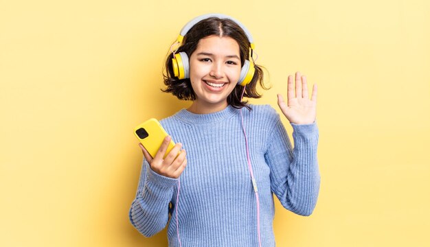 Jovem mulher hispânica sorrindo alegremente, acenando com a mão, dando as boas-vindas e cumprimentando você. conceito de fones de ouvido e smartphone