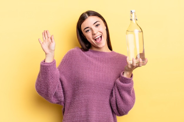 Jovem mulher hispânica sorrindo alegremente, acenando com a mão, dando as boas-vindas e cumprimentando você. conceito de garrafa de água