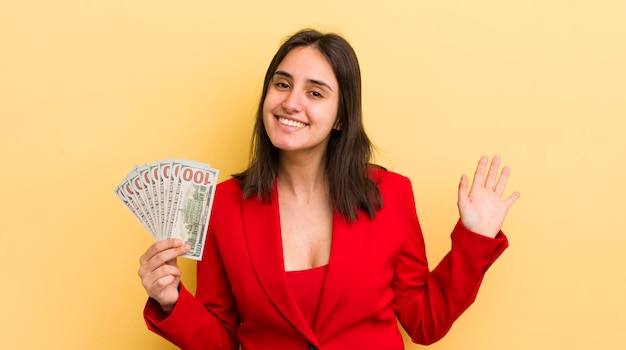 Jovem mulher hispânica sorrindo alegremente acenando com a mão dando as boas-vindas e cumprimentando você conceito de notas de dólar