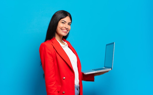 Jovem mulher hispânica sorrindo alegremente com uma mão no quadril e atitude confiante, positiva, orgulhosa e amigável. conceito de laptop