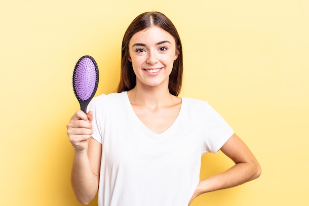 Jovem mulher hispânica sorrindo alegremente com uma mão no quadril e conceito de escova de cabelo confiante