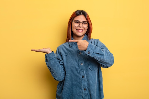 Jovem mulher hispânica sorrindo alegremente e apontando para copiar o espaço na palma da mão mostrando ou anunciando um objeto