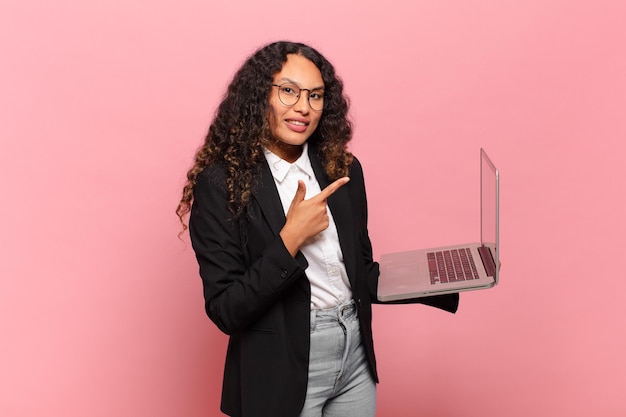 Jovem mulher hispânica sorrindo alegremente, sentindo-se feliz e apontando para os lados e para cima, mostrando o objeto no espaço da cópia. conceito de laptop