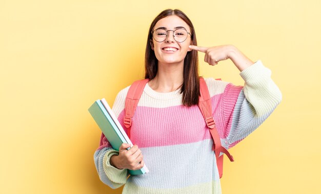 Jovem mulher hispânica sorrindo com confiança, apontando para o próprio sorriso largo. conceito de estudante