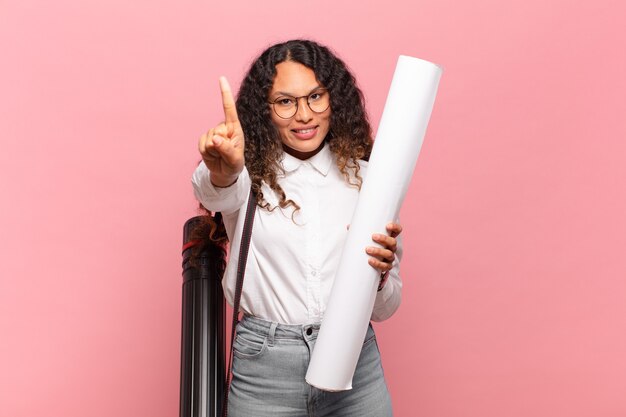 Jovem mulher hispânica sorrindo com orgulho e confiança, fazendo a pose número um triunfantemente, sentindo-se uma líder. conceito de arquiteto