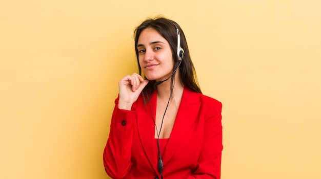 Jovem mulher hispânica sorrindo com uma expressão feliz e confiante com a mão no queixo conceito de telemarketing