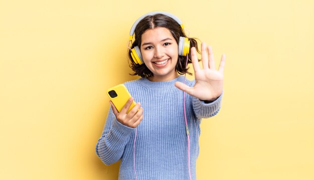 Jovem mulher hispânica sorrindo e parecendo amigável, mostrando o número cinco. conceito de fones de ouvido e smartphone