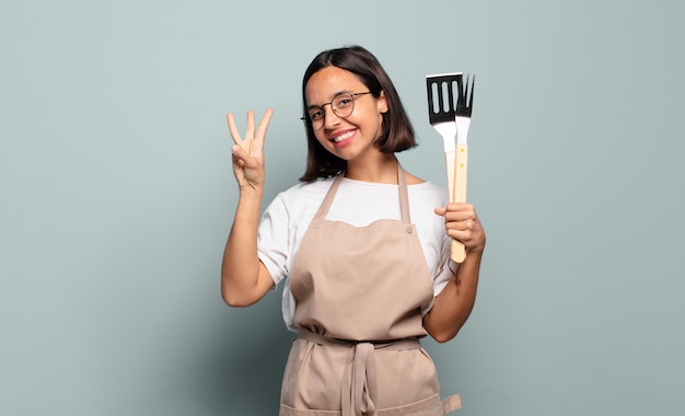 Jovem mulher hispânica sorrindo e parecendo amigável, mostrando o número três ou o terceiro com a mão para a frente, em contagem regressiva