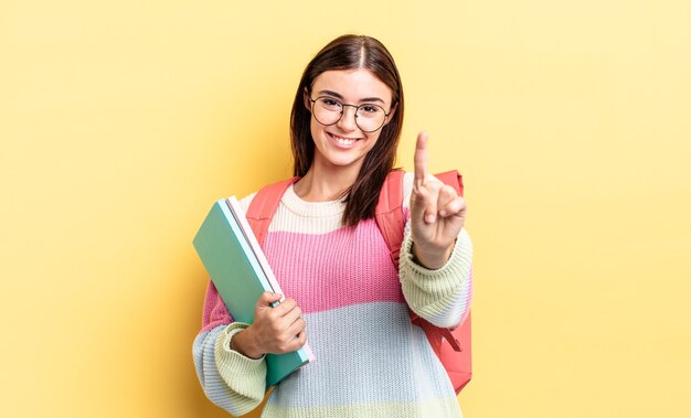 Jovem mulher hispânica sorrindo e parecendo amigável, mostrando o número um. conceito de estudante