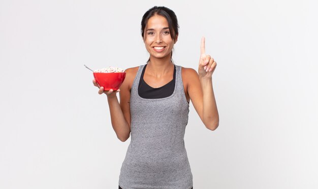 Jovem mulher hispânica sorrindo e parecendo amigável, mostrando o número um. dieta de fitness e conceito de café da manhã