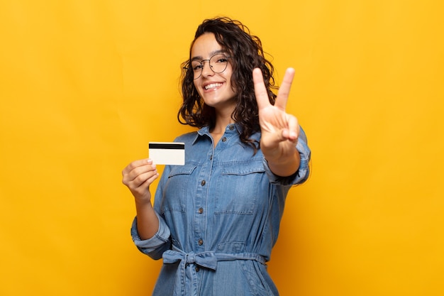 Jovem mulher hispânica sorrindo e parecendo feliz, despreocupada e positiva, gesticulando vitória ou paz com uma mão