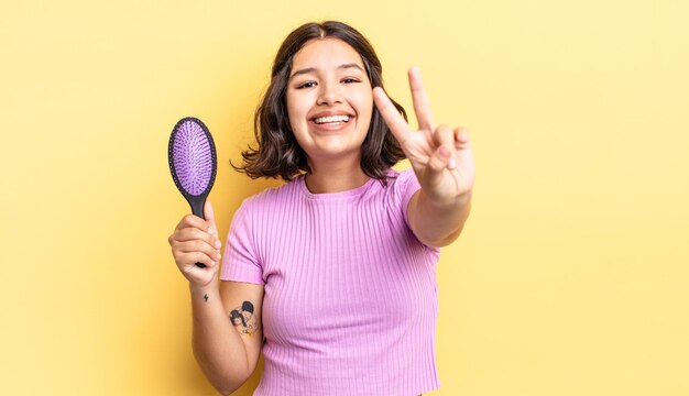 Jovem mulher hispânica sorrindo e parecendo feliz, gesticulando vitória ou paz. conceito de escova de cabelo