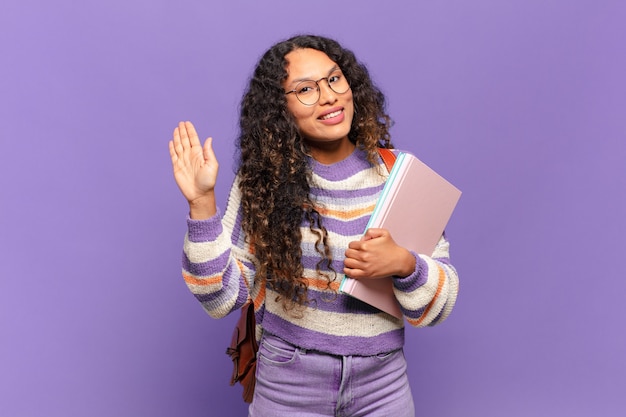 Jovem mulher hispânica sorrindo feliz e alegremente, acenando com a mão, dando as boas-vindas e cumprimentando ou dizendo adeus. conceito de estudante