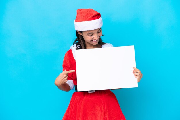 Jovem mulher hispânica vestida como mamãe noel isolada no fundo branco segurando um cartaz vazio com expressão feliz e apontando-o