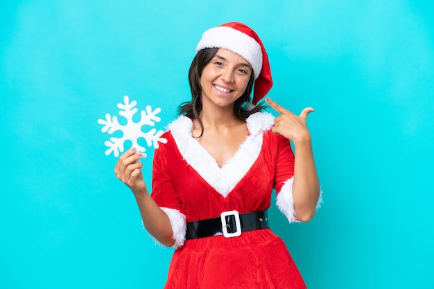 Jovem mulher hispânica vestida como mamãe noel segurando um floco de neve isolado em fundo azul, dando um polegar para cima gesto