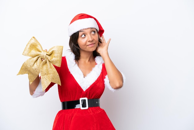 Jovem mulher hispânica vestida como mamãe noel segurando um laço de natal isolado no fundo branco ouvindo algo colocando a mão na orelha