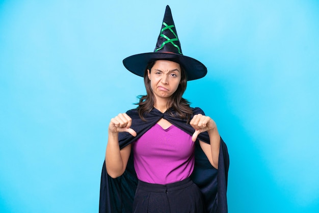 Jovem mulher hispânica vestida de bruxa sobre fundo isolado, mostrando o polegar para baixo com as duas mãos