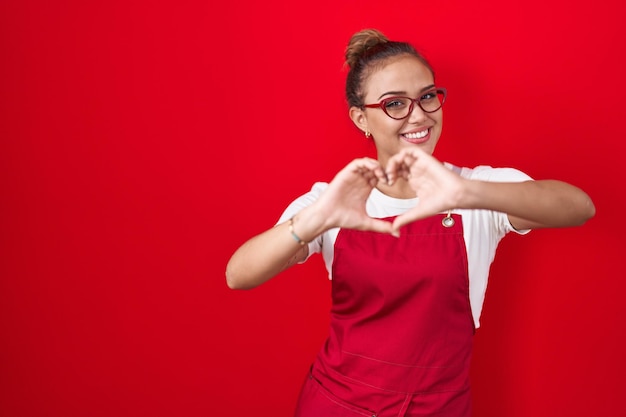 Jovem mulher hispânica vestindo avental de garçonete sobre fundo vermelho sorrindo apaixonadamente fazendo forma de símbolo de coração com as mãos. conceito romântico.