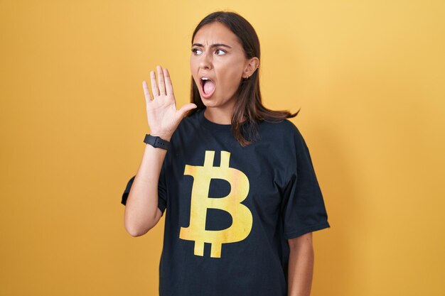 Jovem mulher hispânica vestindo camiseta bitcoin gritando e gritando alto a lado com a mão na boca. conceito de comunicação.