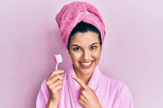 Jovem mulher hispânica vestindo roupão de banho segurando a escova de dentes sorrindo feliz apontando com a mão e o dedo