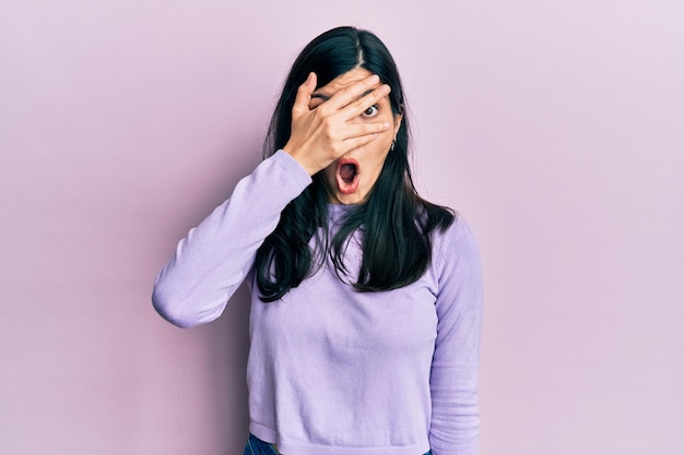 Jovem mulher hispânica vestindo roupas casuais espreitando em estado de choque cobrindo o rosto e os olhos com a mão olhando por entre os dedos com expressão envergonhada