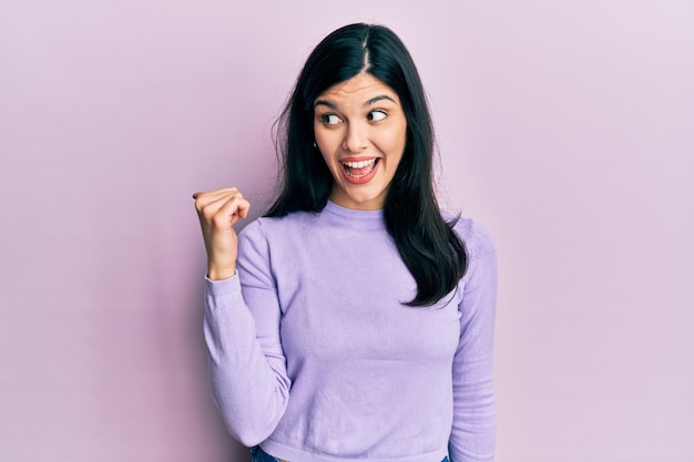 Jovem mulher hispânica vestindo roupas casuais sorrindo com cara de feliz olhando e apontando para o lado com o polegar para cima