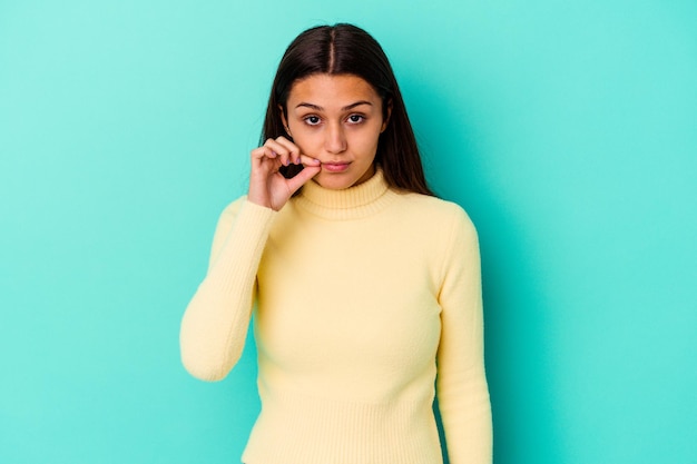 Jovem mulher indiana isolada em azul com os dedos nos lábios, mantendo um segredo.