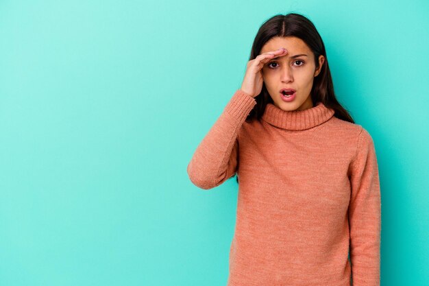 Jovem mulher indiana isolada em azul olhando para longe, mantendo a mão na testa.