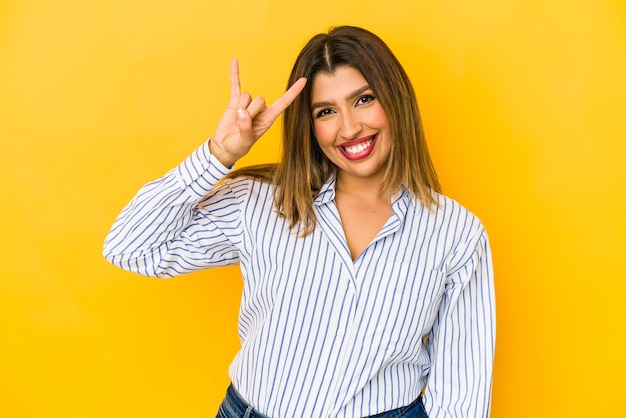 Jovem mulher indiana isolada em fundo amarelo, mostrando um gesto de chifres como um conceito de revolução.