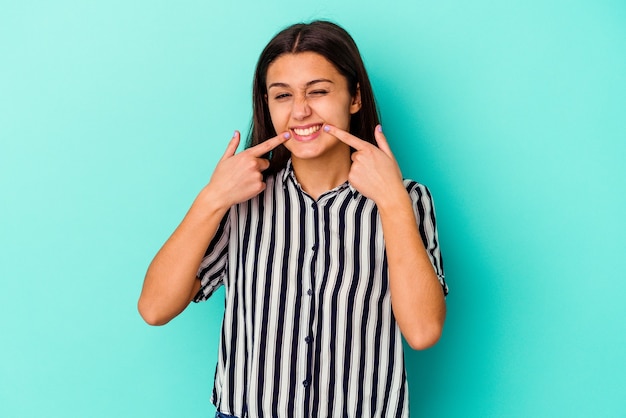 Jovem mulher indiana isolada na parede azul sorri, apontando o dedo para a boca.