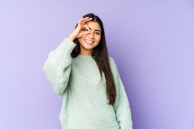 Jovem mulher indiana isolada na parede roxa animada mantendo o gesto ok no olho.