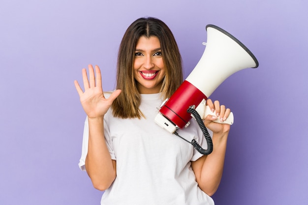 Jovem mulher indiana segurando um megafone isolado sorrindo alegre mostrando o número cinco com os dedos.