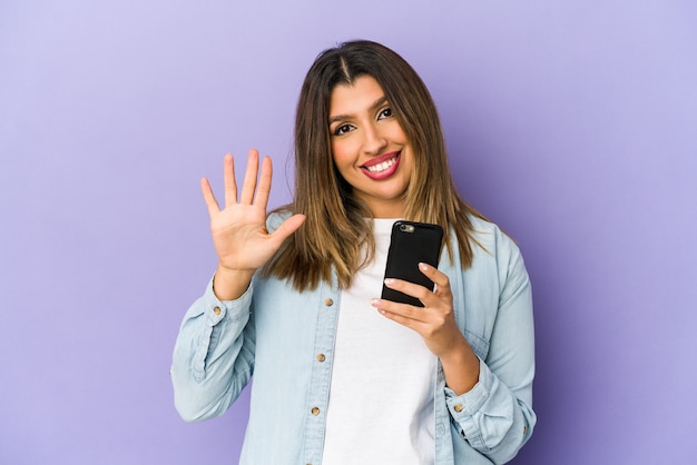 Jovem mulher indiana segurando um telefone isolado sorrindo alegre mostrando o número cinco com os dedos.