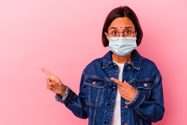 Jovem mulher indiana usando uma máscara antivírus isolada em um fundo rosa chocada apontando com o dedo indicador para um espaço de cópia.