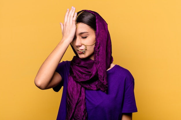 Jovem mulher indiana vestindo uma roupa de sari tradicional isolada na parede amarela, esquecendo algo, batendo na testa com a palma da mão e fechando os olhos.