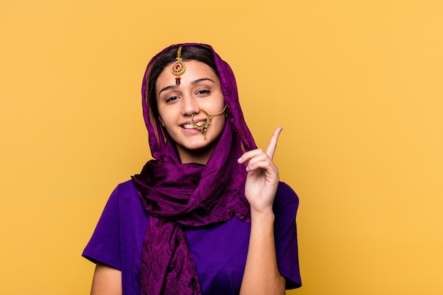 Jovem mulher indiana vestindo uma roupa de sari tradicional, isolada na parede amarela, sorrindo alegremente, apontando com o dedo indicador para longe.